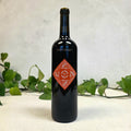 Black wine bottle with a red label on a white surface with green leaves in the background