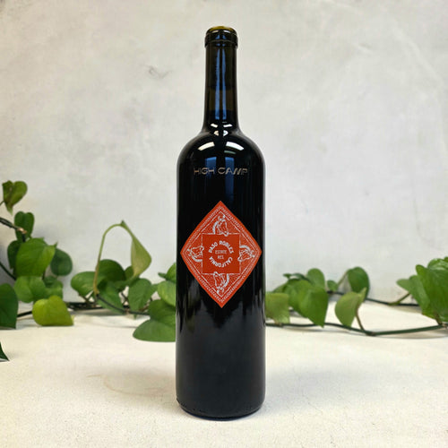 Black wine bottle with a red label on a white surface with green leaves in the background