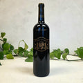 Black wine bottle with decorative label on a light background with green leaves.
